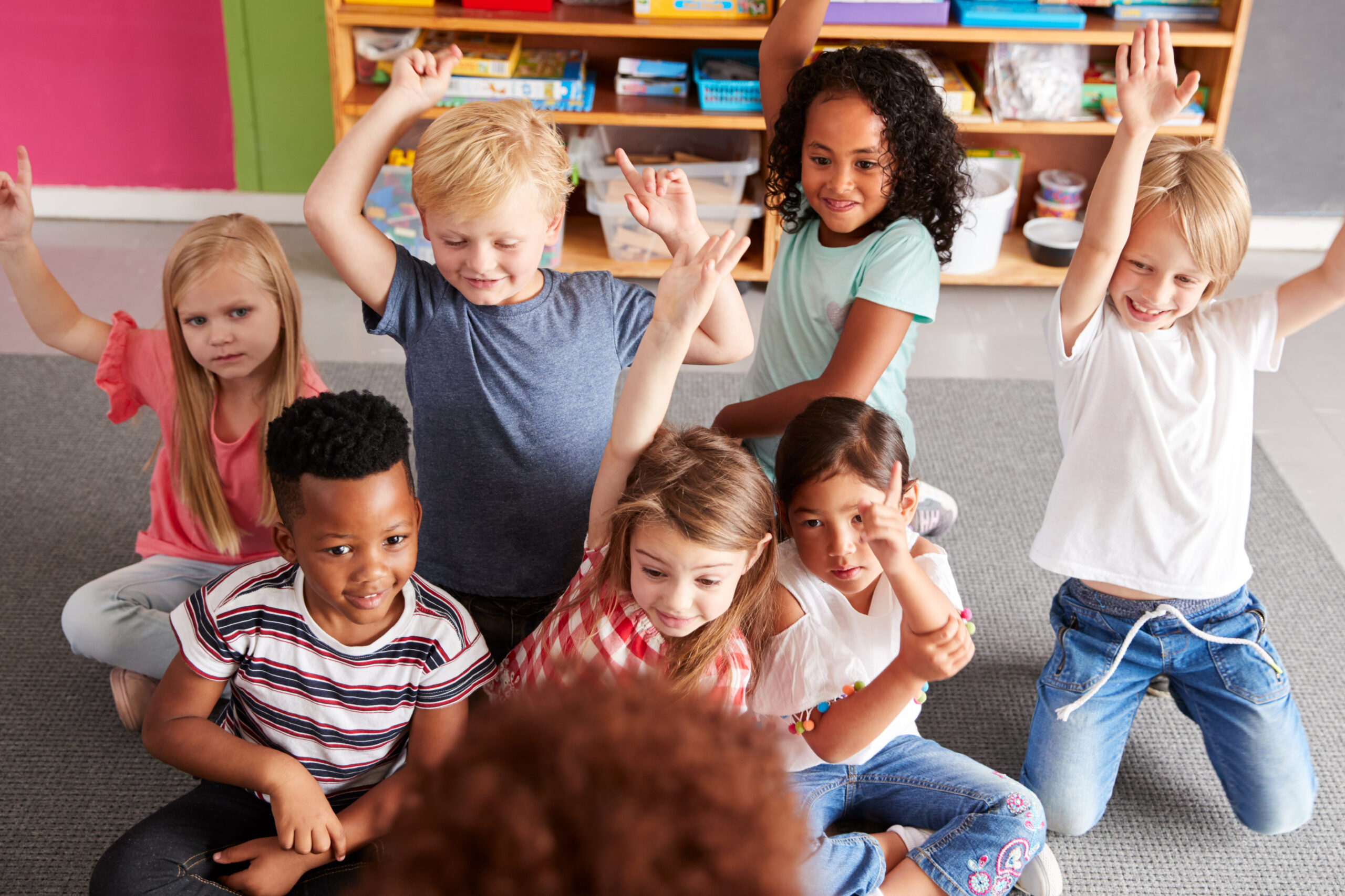 Elementary Pupils Raising Hands To Answer Question As Female Teacher Reads Story In Classroom